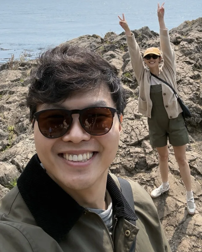 A selfie of Jason on a rocky shoreline. Jason's wife waves in the background.